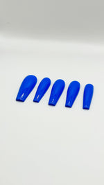 A top-down product photograph showing a set of five long, high-gloss royal blue press-on nails arranged neatly in a row. The salon-grade gel nails are displayed on a clean, solid white background, showcasing their crisp shape, rich solid blue color, and a flawless, reflective glass-like top coat.