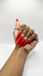 A close-up of a hand wearing long, tapered square press-on nails in a solid, vibrant cherry red color with a high-gloss finish. The nails are plain with no added charms or art. The hand is styled with multiple gold rings against a white background.