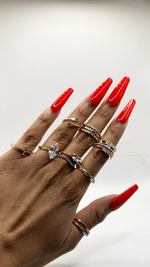 A close-up of a hand with long, glossy red manicured nails wearing multiple stacked rings, including a heart-shaped diamond ring and textured gold bands, against a plain light background.