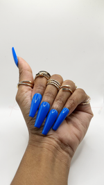 A hand with long, glossy bright blue square-shaped press-on nails, accessorized with multiple gold rings, shown against a plain white background.