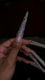 A close-up side view of a hand highlighting an extra-long stiletto press-on thumbnail. The nail features a shimmering iridescent pink base with the entire length of the extended tip fully encrusted in textured, sparkling clear pixie crystals and rhinestones.