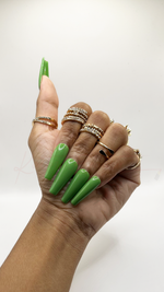 A hand with long, glossy green coffin-shaped press-on nails, accessorized with multiple gold rings, shown against a plain white background.