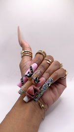Extra-long, square press-on nails with a pink and nude color palette. The index finger is light bubblegum pink, wrapped diagonally in a black rhinestone and silver-beaded BB belt design. The middle finger has a nude base featuring a 3D bikini design with black bikini cups shaped like cartoon character faces. The ring finger has a glossy pink base dominated by a large, 3D hand-sculpted open mouth charm with pink lips. The pinky finger is covered from base to tip in shimmering rhinestones