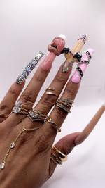 A hand with fingers spread straight open, wearing numerous gold rings and a finger chain bracelet, showcasing an extra-long, square press-on nail set. The index nail is pink with a diagonal black rhinestone BB belt design. The middle nail is nude with a 3D bikini design featuring black cartoon character bikini. The ring nail is pink with a large 3D hand-sculpted mouth charm with a black grill. The pinky nail is fully encrusted with shimmering iridescent rhinestones.