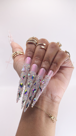 A close-up of a hand wearing extra-long stiletto press-on nails. Features a shimmering iridescent pink base with the entire length of the extended tips fully encrusted in clear pixie crystals. Accentuated with scattered iridescent diamond-cut rhinestones.
