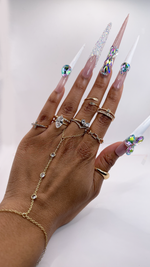 A close-up of a hand wearing extra-long stiletto press-on nails. Features a glossy nude base with clean white French tips. Large iridescent teardrop rhinestones are placed on the nails, with one accent nail fully covered in textured pixie crystals.