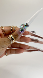 A close-up side view of a hand highlighting an extra-long stiletto press-on thumbnail. The nail features a glossy nude base with a crisp white French tip. A cluster of three large iridescent, multi-faceted teardrop rhinestones sits where the nude base meets the white tip.