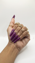A hand with long, glossy purple coffin-shaped press-on nails, styled with multiple gold rings, shown against a plain white background.
