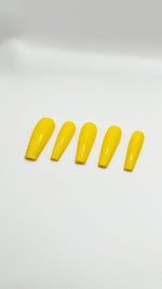 A top-down product photograph showing a set of five long, high-gloss banana yellow press-on nails arranged neatly in a row. The salon-grade gel nails are displayed on a clean, solid white background, highlighting their crisp shape, uniform solid color, and flawless glass-like reflective top coat.