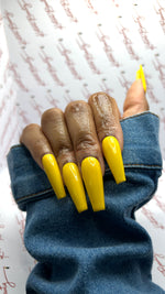 A close-up photograph of a hand wearing long, glossy, vibrant yellow coffin-shaped press-on nails. The fingers are lightly moisturized with cuticle oil, highlighting the flawless, clean application at the cuticle line. The hand is pulling up the sleeve of a classic blue denim jacket against a bright, professional branding background.