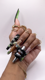 A close-up of a hand wearing extra-long, square-shaped press-on nails with complex, multi-textured 3D art. The color palette features neon lime green, jet black, shiny silver foil, and nude. One nail has a large, glossy black 3D charm shaped like open lips showing teeth with tiny rhinestones inside. Another nail features a massive, iridescent 3D butterfly charm sitting over a green and black marbled pattern. Other nails are decorated with wrapped silver rhinestone chains, metallic silver textures, and clear