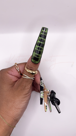 Side view of an extra long square press-on thumbnail features a realistic 3D crocodile skin leather texture with glossy, raised black and deep olive green ridges. Hand is styled with gold rings on a plain white background.