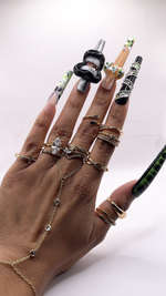 A close-up of a hand wearing extra-long square press-on nails with 3D art. Features a glossy black lip charm with teeth, an iridescent butterfly piece, wrapped rhinestone chains, green marbling, and a crocodile skin thumb. Wears gold hand jewelry.
